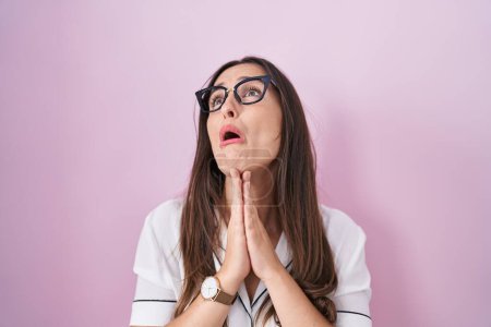 Photo for Young brunette woman wearing glasses standing over pink background begging and praying with hands together with hope expression on face very emotional and worried. begging. - Royalty Free Image