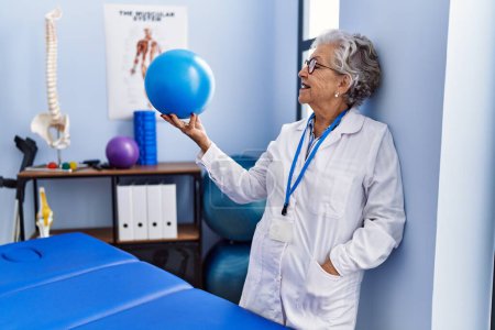 Foto de Mujer mayor de cabello gris con uniforme de fisioterapeuta sosteniendo la pelota en la clínica de fisioterapia - Imagen libre de derechos