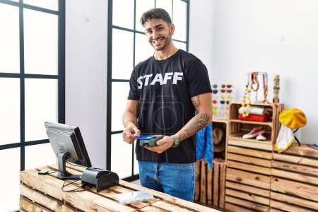 Foto de Hombre hispano guapo trabajando como asistente de tienda tomando el pago de la tarjeta en la tienda al por menor - Imagen libre de derechos