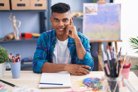 Téléchargez les photos : Jeune homme latin artiste souriant dessin confiant sur carnet à l'atelier d'art - en image libre de droit