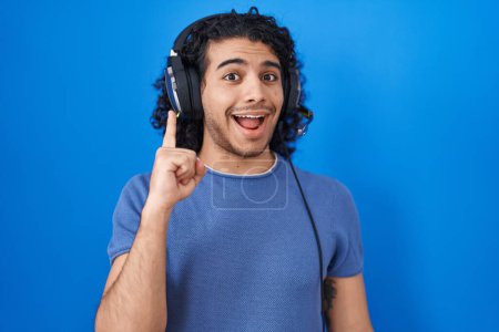 Téléchargez les photos : Hispanic man with curly hair listening to music using headphones pointing finger up with successful idea. exited and happy. number one. - en image libre de droit