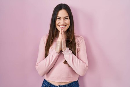 Photo for Young brunette woman standing over pink background praying with hands together asking for forgiveness smiling confident. - Royalty Free Image