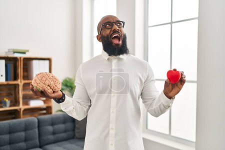 Foto de African american man working at therapy office holding brain and heart angry and mad screaming frustrated and furious, shouting with anger looking up. - Imagen libre de derechos
