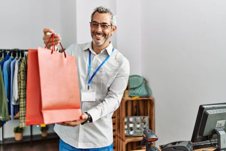 Téléchargez les photos : Moyen-âge homme aux cheveux gris boutique assistant tenant sac à provisions au magasin de vêtements - en image libre de droit