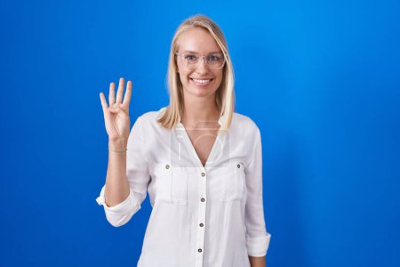 Téléchargez les photos : Jeune femme caucasienne debout sur fond bleu montrant et pointant vers le haut avec les doigts numéro quatre tout en souriant confiant et heureux. - en image libre de droit