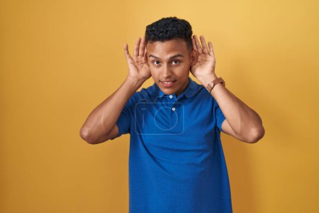 Téléchargez les photos : Jeune homme hispanique debout sur fond jaune essayant d'entendre les deux mains sur le geste de l'oreille, curieux de commérages. problème auditif, sourd - en image libre de droit