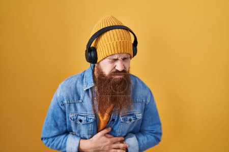 Photo for Caucasian man with long beard listening to music using headphones with hand on stomach because indigestion, painful illness feeling unwell. ache concept. - Royalty Free Image