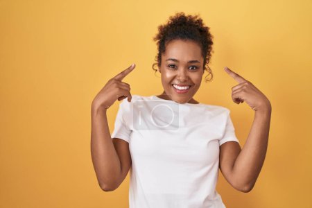 Photo for Young hispanic woman with curly hair standing over yellow background smiling pointing to head with both hands finger, great idea or thought, good memory - Royalty Free Image
