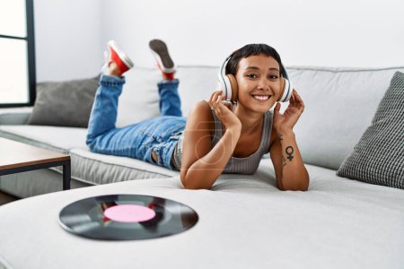 Foto de Mujer hispana joven escuchando música tumbada en un sofá en casa - Imagen libre de derechos