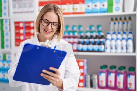 Téléchargez les photos : Young blonde woman pharmacist smiling confident writing on document at pharmacy - en image libre de droit