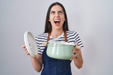 Téléchargez les photos : Young brunette woman wearing apron holding cooking pot angry and mad screaming frustrated and furious, shouting with anger looking up. - en image libre de droit