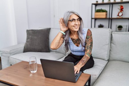 Photo for Middle age grey-haired woman using laptop at home smiling with hand over ear listening an hearing to rumor or gossip. deafness concept. - Royalty Free Image