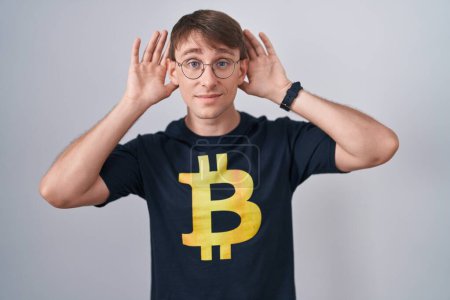 Photo for Caucasian blond man wearing bitcoin t shirt trying to hear both hands on ear gesture, curious for gossip. hearing problem, deaf - Royalty Free Image