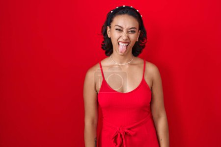 Foto de Young hispanic woman standing over red background sticking tongue out happy with funny expression. emotion concept. - Imagen libre de derechos