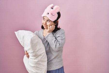 Foto de Woman with down syndrome wearing sleeping mask hugging pillow looking stressed and nervous with hands on mouth biting nails. anxiety problem. - Imagen libre de derechos