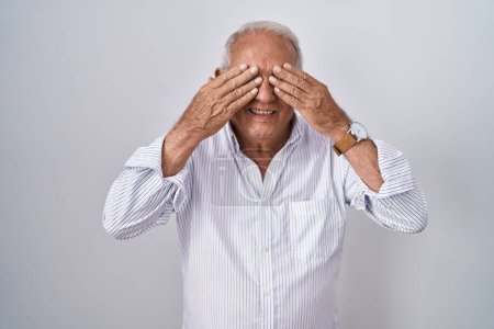 Foto de Senior man with grey hair standing over isolated background covering eyes with hands smiling cheerful and funny. blind concept. - Imagen libre de derechos