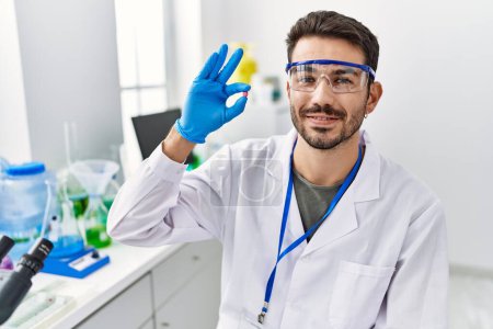 Téléchargez les photos : Young hispanic man wearing scientist uniform holding pill at laboratory - en image libre de droit