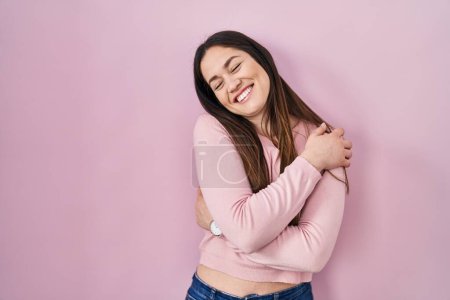 Foto de Joven morena de pie sobre fondo rosa abrazándose feliz y positivo, sonriendo confiado. amor propio y cuidado personal - Imagen libre de derechos