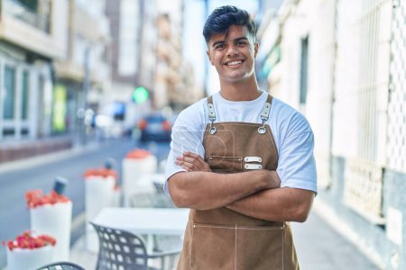 Foto de Joven camarero hispano sonriendo confiado de pie con los brazos cruzados gesto en la terraza de la cafetería - Imagen libre de derechos