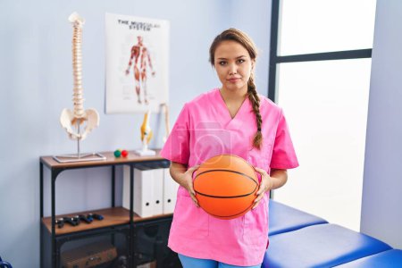 Foto de Young hispanic woman working at physiotherapy clinic holding basketball ball relaxed with serious expression on face. simple and natural looking at the camera. - Imagen libre de derechos