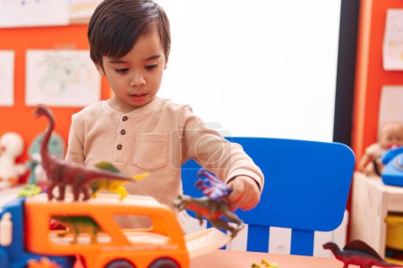 Photo for Adorable hispanic boy playing with cars and dino toys standing at kindergarten - Royalty Free Image