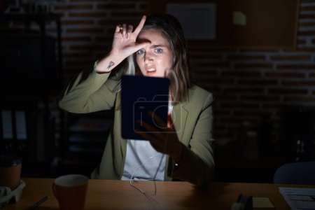 Photo for Blonde caucasian woman working at the office at night making fun of people with fingers on forehead doing loser gesture mocking and insulting. - Royalty Free Image
