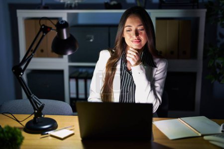 Foto de Young brunette woman working at the office at night with laptop looking confident at the camera smiling with crossed arms and hand raised on chin. thinking positive. - Imagen libre de derechos