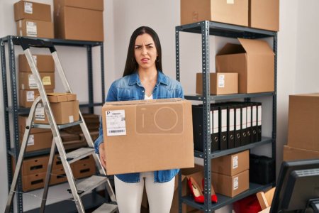 Foto de Hispanic woman working at small business ecommerce holding box clueless and confused expression. doubt concept. - Imagen libre de derechos