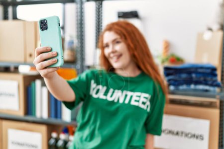Foto de Joven pelirroja vistiendo uniforme voluntario hacer selfie por el teléfono inteligente en el centro de caridad - Imagen libre de derechos