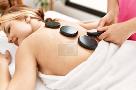 Téléchargez les photos : Jeune femme caucasienne allongée sur la table ayant massage du dos en utilisant des pierres chaudes au salon de beauté - en image libre de droit
