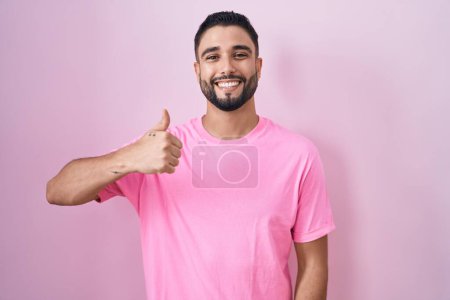 Photo for Hispanic young man standing over pink background doing happy thumbs up gesture with hand. approving expression looking at the camera showing success. - Royalty Free Image