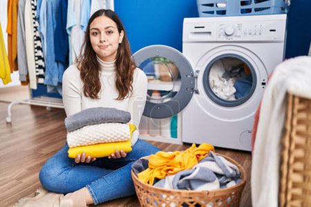Téléchargez les photos : Young hispanic woman holding clean laundry puffing cheeks with funny face. mouth inflated with air, catching air. - en image libre de droit