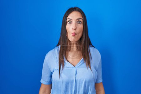 Photo for Young brunette woman standing over blue background making fish face with lips, crazy and comical gesture. funny expression. - Royalty Free Image