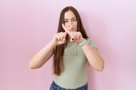 Téléchargez les photos : Beautiful brunette woman standing over pink background rejection expression crossing fingers doing negative sign - en image libre de droit