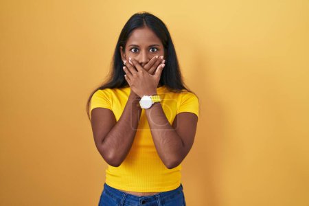 Foto de Young indian woman standing over yellow background shocked covering mouth with hands for mistake. secret concept. - Imagen libre de derechos