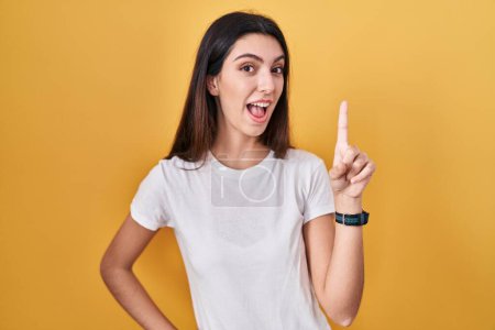 Foto de Young beautiful woman standing over yellow background pointing finger up with successful idea. exited and happy. number one. - Imagen libre de derechos
