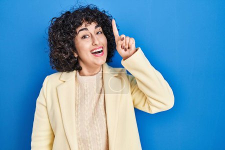 Photo for Young brunette woman with curly hair standing over blue background pointing finger up with successful idea. exited and happy. number one. - Royalty Free Image
