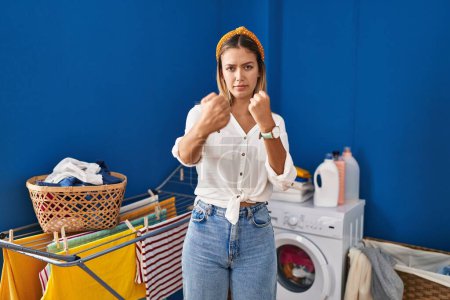 Photo for Young blonde woman at laundry room ready to fight with fist defense gesture, angry and upset face, afraid of problem - Royalty Free Image