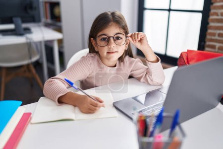 Photo for Adorable hispanic girl student using laptop writing on notebook at classroom - Royalty Free Image