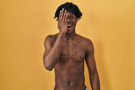 Foto de Young african man with dreadlocks standing shirtless covering one eye with hand, confident smile on face and surprise emotion. - Imagen libre de derechos