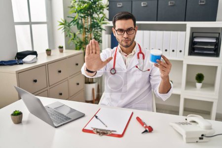 Foto de Young hispanic doctor man with beard holding ear cotton buds with open hand doing stop sign with serious and confident expression, defense gesture - Imagen libre de derechos