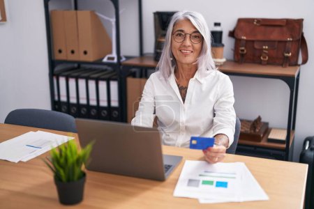 Téléchargez les photos : Femme d'âge moyen aux cheveux gris qui travaille avec un ordinateur portable et une carte de crédit au bureau - en image libre de droit