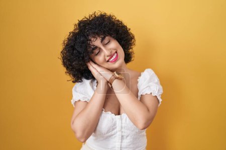 Foto de Young brunette woman with curly hair standing over yellow background sleeping tired dreaming and posing with hands together while smiling with closed eyes. - Imagen libre de derechos