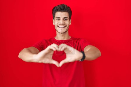 Foto de Young hispanic man standing over red background smiling in love doing heart symbol shape with hands. romantic concept. - Imagen libre de derechos