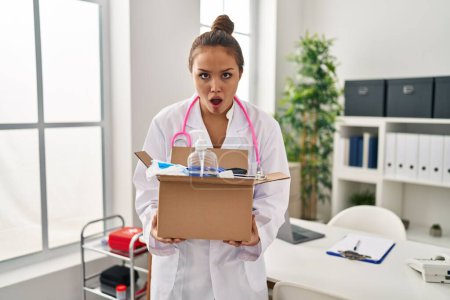 Téléchargez les photos : Young hispanic doctor holding box with medical items in shock face, looking skeptical and sarcastic, surprised with open mouth - en image libre de droit