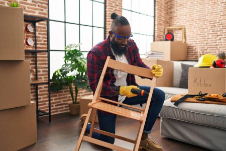 Téléchargez les photos : Jeune homme afro-américain ouvrier réparation chaise à la nouvelle maison - en image libre de droit