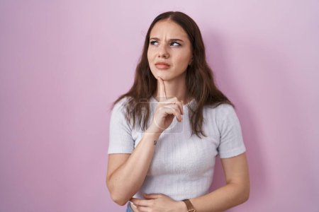 Photo for Young hispanic girl standing over pink background thinking concentrated about doubt with finger on chin and looking up wondering - Royalty Free Image