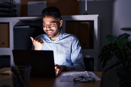 Photo for Hispanic man with beard working at the office with laptop at night pointing thumb up to the side smiling happy with open mouth - Royalty Free Image