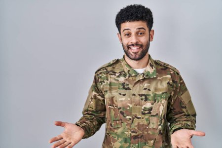 Foto de Hombre árabe vistiendo uniforme camuflaje ejército sonriente alegre con los brazos abiertos como bienvenida amistosa, saludos positivos y confiados - Imagen libre de derechos