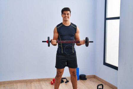 Téléchargez les photos : Jeune homme hispanique souriant confiant en utilisant bar avec entraînement de disque au centre sportif - en image libre de droit